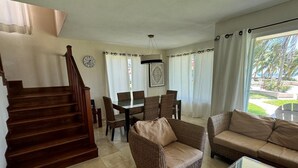 Een ruime woonkamer met een houten trap, rieten meubels en een eettafel met stoelen.