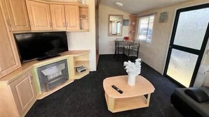 Een compact woongedeelte met een flatscreen televisie op een houten kast, een klein houten tafeltje met een vaas, een zwarte bank en een eethoek met een glazen tafel en stoelen.