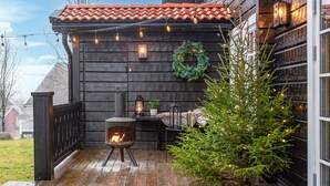 Una terraza de madera con un estufa de leña, una pequeña mesa con faroles y un árbol de Navidad.