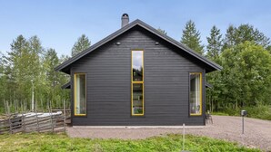 Une cabane moderne en bois sombre, dotée de deux grandes fenêtres et d’une cheminée, et entourée d’un cadre forestier naturel.