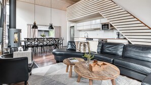 Un salon moderne avec un canapé en cuir noir, une table basse en bois et un coin repas comprenant une table et des chaises en bois. On aperçoit au fond une cuisine avec des meubles blancs et un four à micro-ondes.