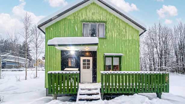 Een groene houten hut met een veranda, een deur en een klein raam.