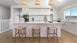 A modern kitchen with white cabinetry, a central island with stools, pendant lighting, and a view through a large window. - Ruby Jade Coastal Luxury at Anna Bay (Anna Bay)