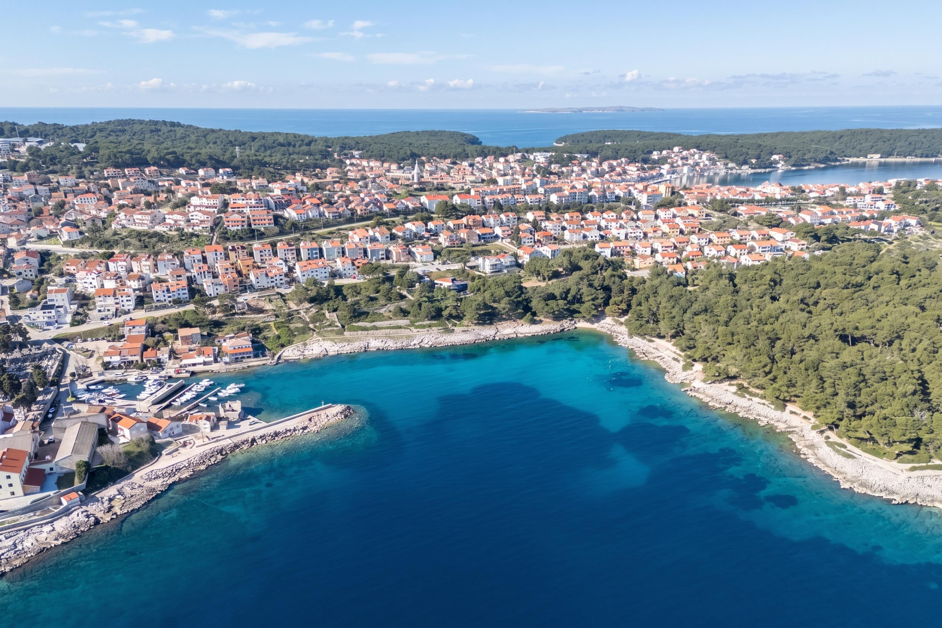 An aerial view of a coastal town with a marina, dense residential area, and a clear blue sea.