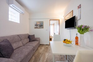 Uma sala de estar moderna com um sofá cinzento, uma mesa branca com uma fruteira, uma TV de tela plana e um quadro na parede.