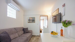 En stue med en sofa, et spisebord med frugt i en skål, et fjernsyn og et billede på væggen.