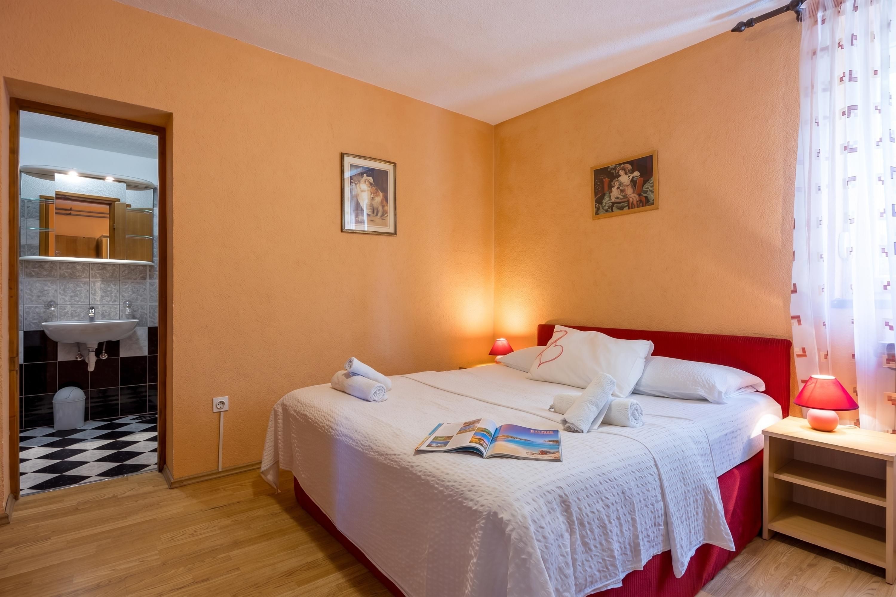 Ein Hotelzimmer mit Bett, Nachttisch, Lampe, einem Fenster mit Vorhängen, einem Wandbild und einem durchgehenden Türspalt sichtbaren Badezimmer.