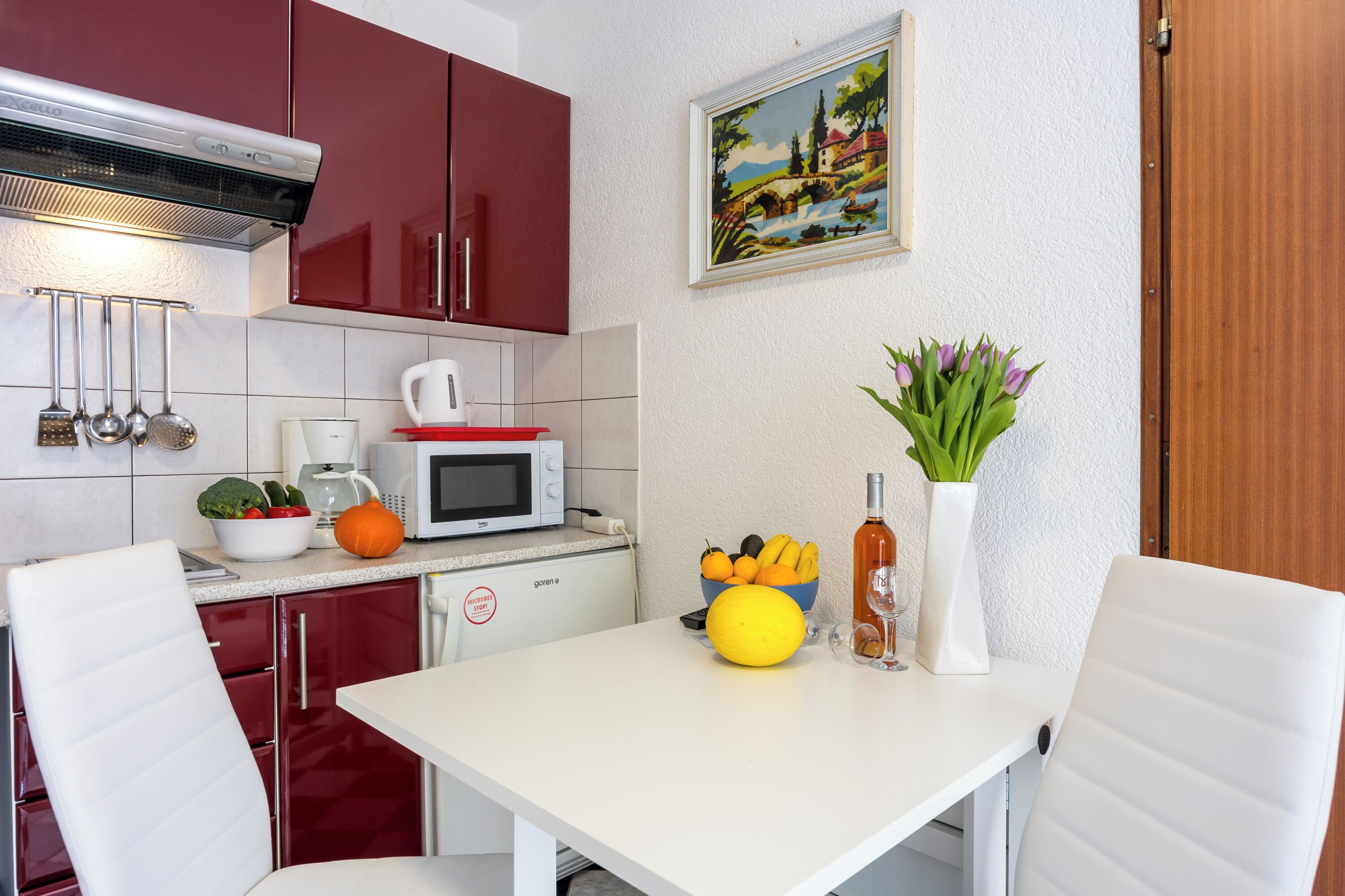 A kitchen with red cabinets, a white countertop, a microwave, a coffee maker, and a table with fruits and a vase.