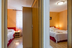 A hotel room with two beds, a nightstand with a lamp, a window with curtains, and a mirror reflecting the room.