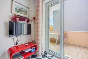 Habitación con una televisión de pantalla plana montada en la pared, un mueble de entretenimiento rojo con libros y un ventanal corredizo que da a un balcón exterior con mesa y sillas.