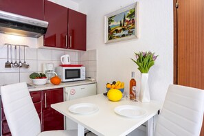 A compact kitchen with red cabinets, a white countertop, a microwave, a kettle, and a dining area with a white table and chairs. - Amalija Sea View Apartment 1, Mali Lošinj, Croatia (Mali Lošinj)