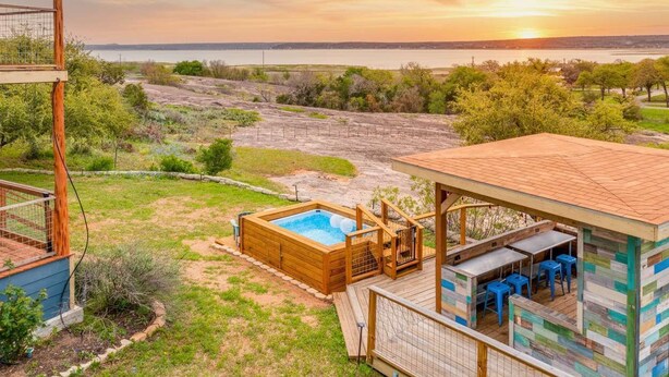 Una terraza de madera con un jacuzzi, asientos exteriores y vistas a la puesta de sol sobre el agua.