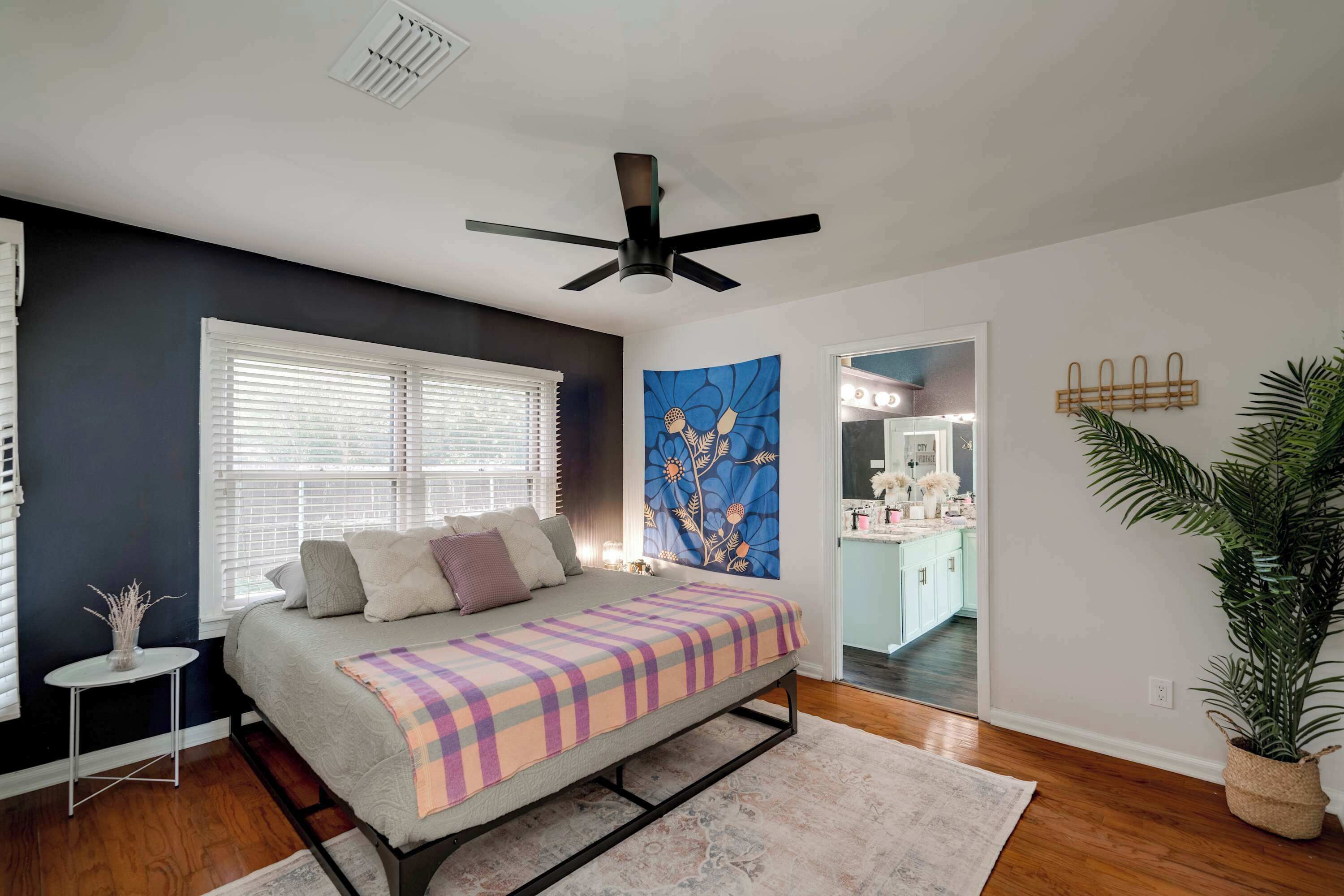 Quarto com cama, ventilador de teto, janela com persianas, um quadro e vista para a cozinha.