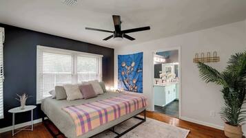 Quarto com cama, ventilador de teto, janela com persianas, um quadro e vista para a cozinha.