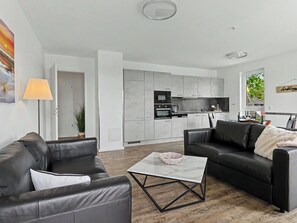 Ein modernes Wohnzimmer mit Ledersofas, einem Glastisch und einer Küche mit grauen Schränken und Geräten.