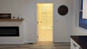 Une pièce avec un plancher en bois, une cheminée, une horloge au mur et une porte donnant sur une salle de bain.