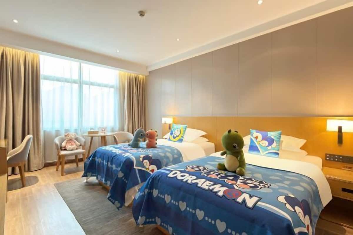 A hotel room with two beds, each with a blue bedspread featuring cartoon characters and the name 'Doraemon.' There are plush toys on the beds, a small table with a lamp, and a chair.
