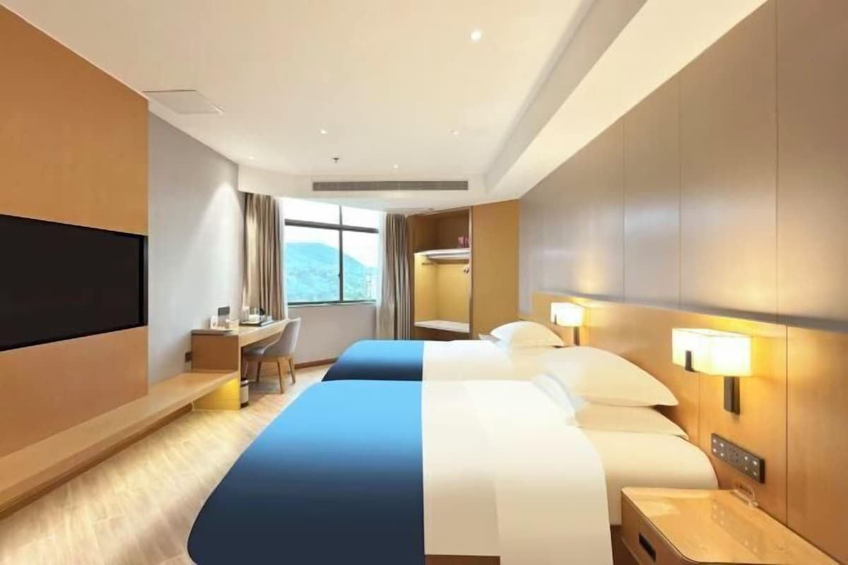 Una habitación de hotel con cama, mesitas de noche, escritorio, televisor y ventana con cortinas.