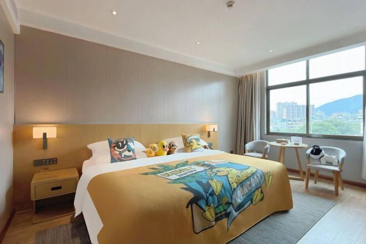 A hotel room with a large bed, a nightstand, a chair, a small table, and a window with a city view.