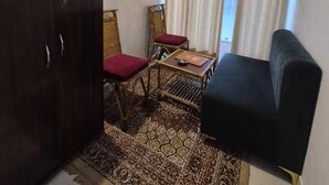 A small living area with a dark-colored sofa, a wooden table with two chairs, a patterned rug, a dark wooden cabinet, and a window with curtains. - Selfie Point Hotel & Restaurant Cafe (Jakhnidhar)