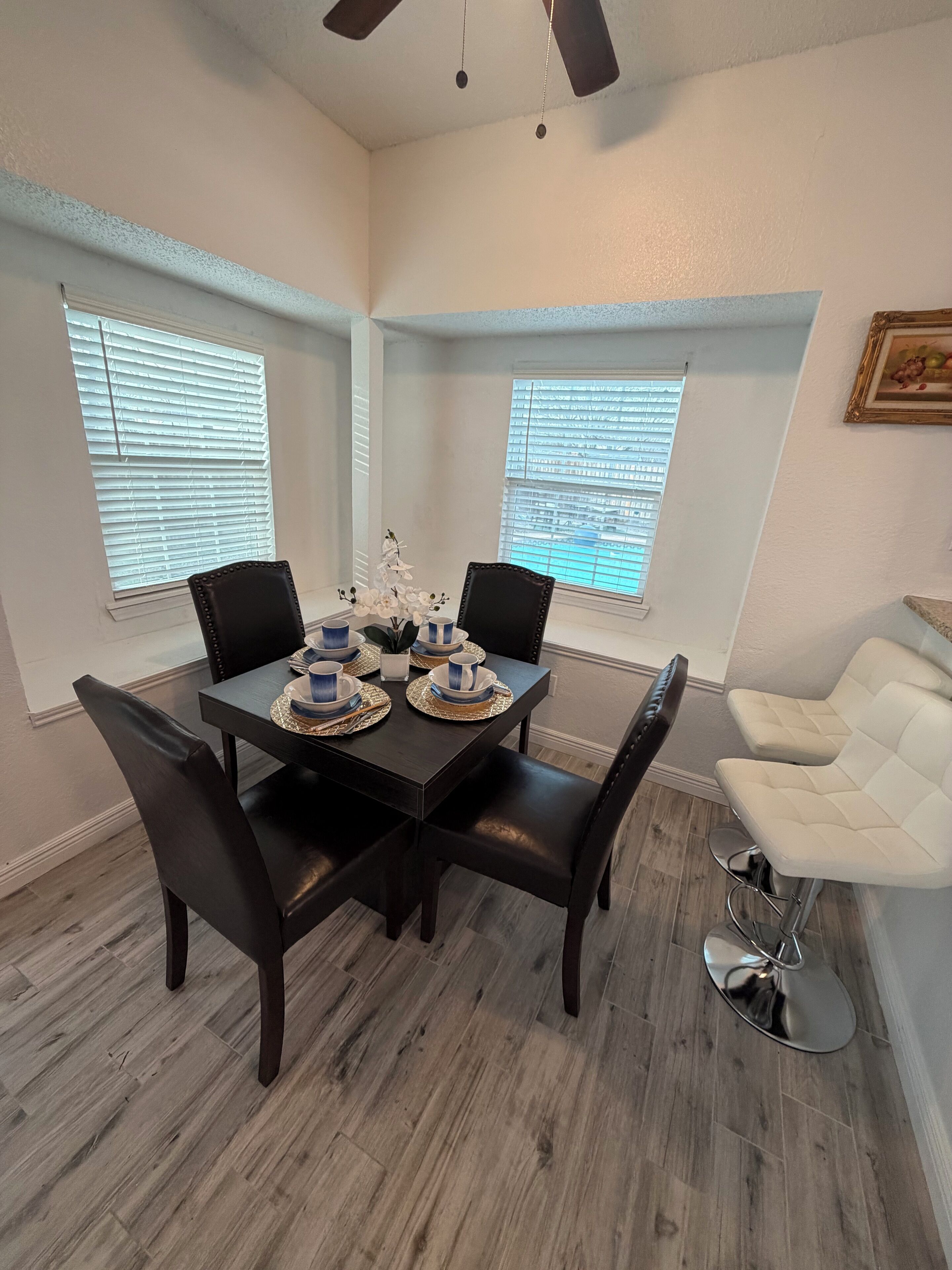 A dining area with a dark wooden table set for four, flanked by black chairs. There are two windows with blinds, a ceiling fan, and a white chair to the right.