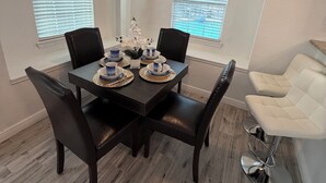 A dining area with a dark wooden table set for four, flanked by black chairs. There are two windows with blinds, a ceiling fan, and a white chair to the right.