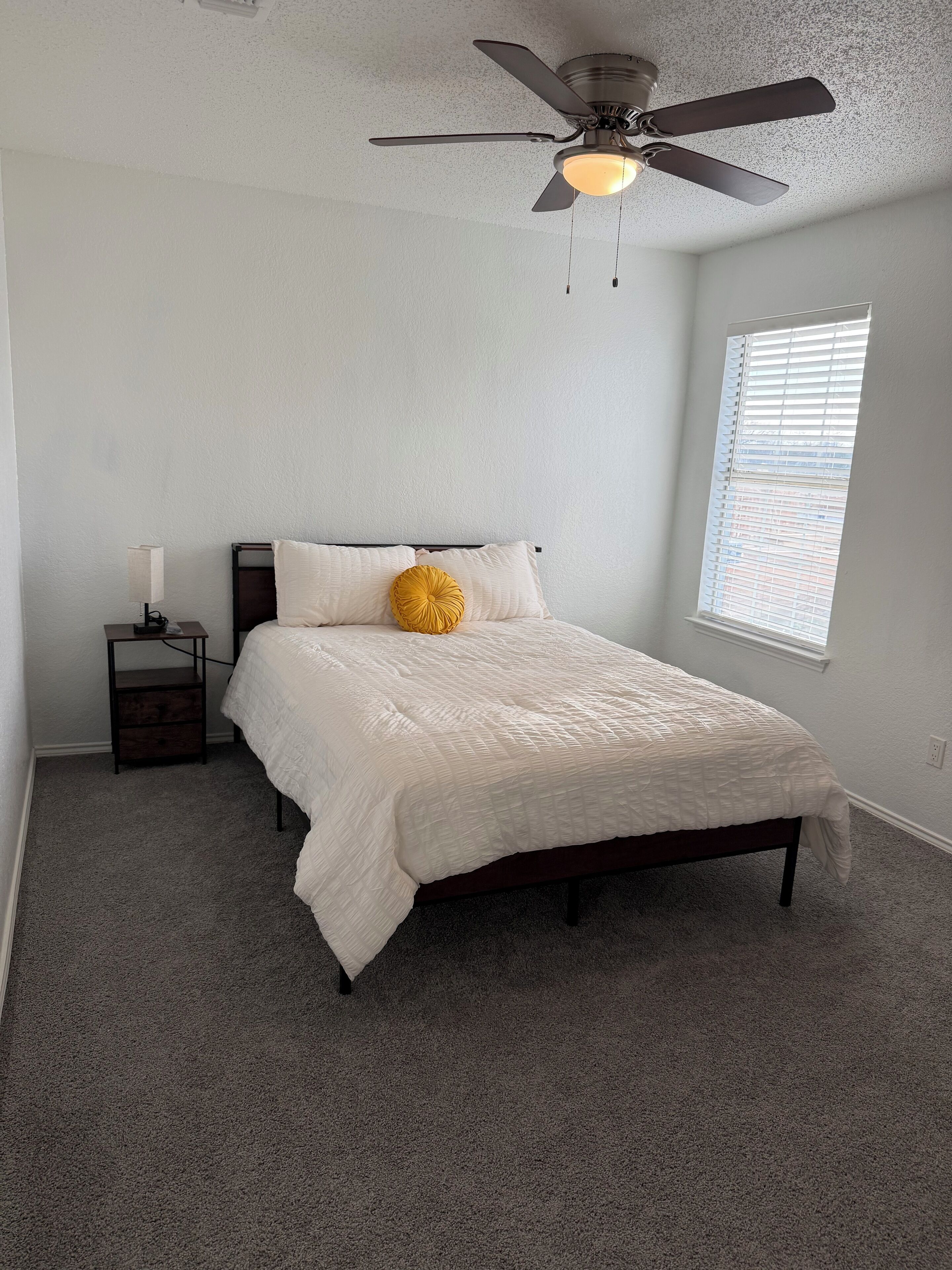 Une chambre à coucher comprenant un lit, une table de chevet, une lampe, un ventilateur de plafond et une fenêtre avec des stores.