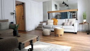 Ein modernes Wohnzimmer mit einer Couch, einem Hocker und einem Couchtisch. Im Hintergrund befindet sich eine Holztür und eine Treppe.