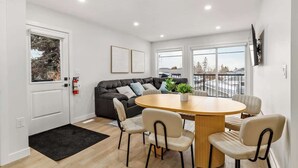 A modern living room with a round wooden table, four chairs, a black leather sofa, and a large window. - AC, 95 min Banff, Local Coffee,Walk to Restaurants (Calgary)