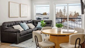 A modern living room with a dark leather sectional sofa, a wooden dining table with four chairs, and large windows offering a view of the outside. - AC, 95 min Banff, Local Coffee,Walk to Restaurants (Calgary)
