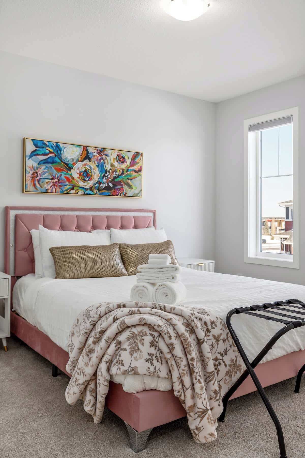 A bedroom with a pink upholstered headboard, a large floral painting above the bed, and a window with a view of buildings outside.