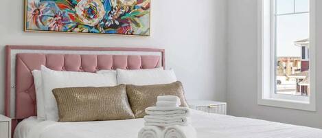 A bedroom with a pink upholstered headboard, a large floral painting above the bed, and a window with a view of buildings outside.