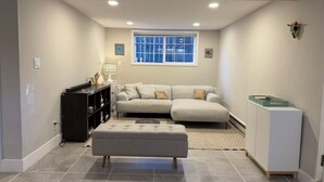 Una sala de estar con un sofá modular, un otoman gris, un aparador de madera oscura y un mueble blanco.