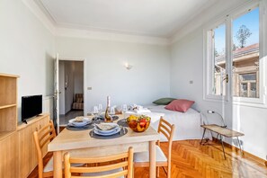Una stanza con tavolo in legno apparecchiato per due, un letto con cuscino verde, una finestra con vista sugli edifici e una televisione su un mobile in legno.