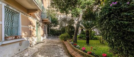 A stone-paved courtyard with a brick flower bed, a building with balconies, and lush greenery.