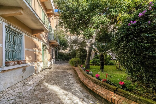 Une cour dallée de pierres, avec un parterre fleuri en briques, un bâtiment doté de balcons et une végétation luxuriante.