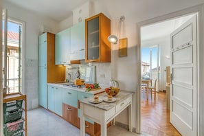 A compact kitchen with teal cabinets, a wooden range hood, and a dining area visible through an open door. - Appartamento Villa Mary (Sanremo)