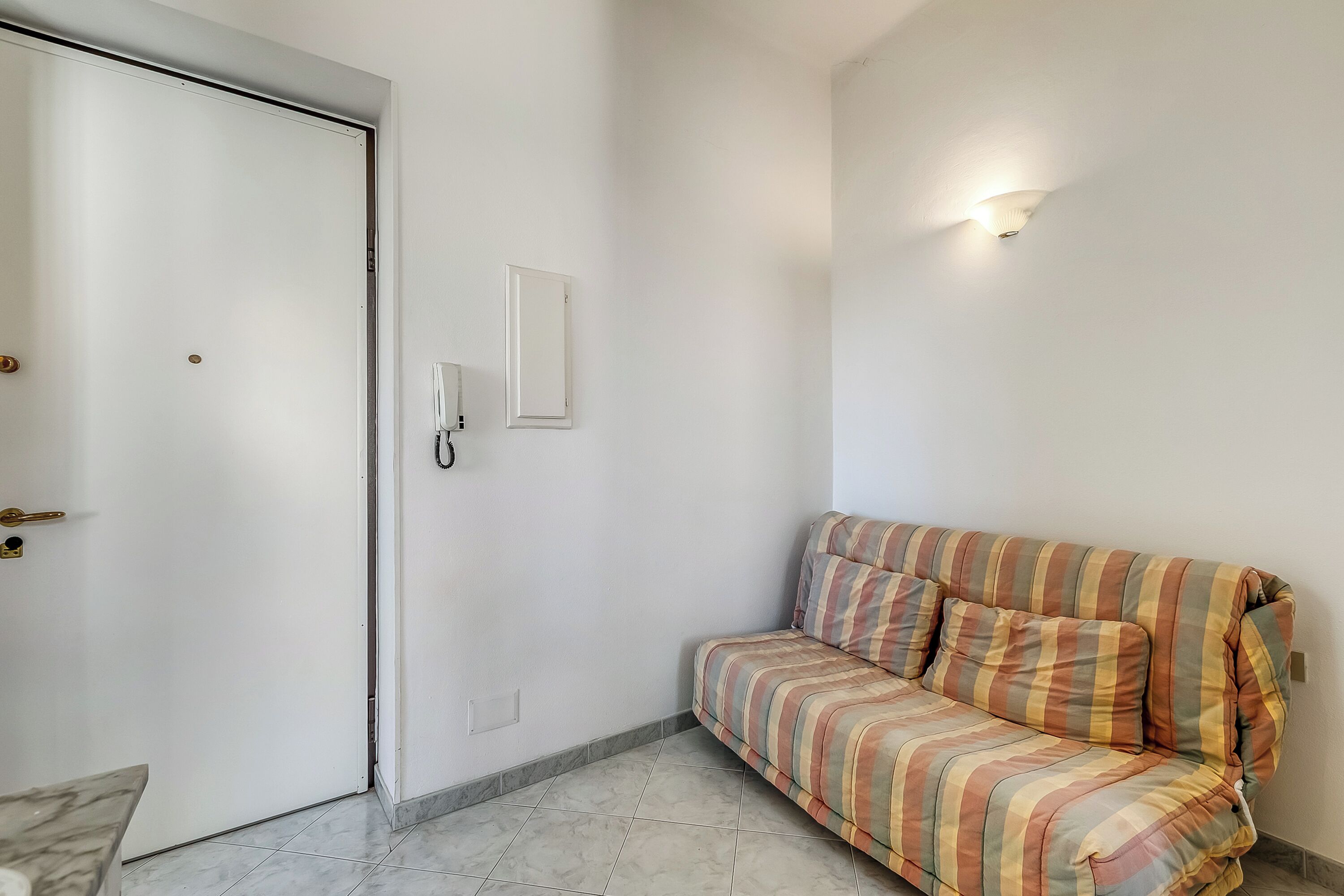 Una habitación con un sofá a rayas, una puerta cerrada y una lámpara fijada a la pared.