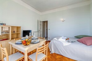 A room with a wooden dining table set for two, a bed with a green pillow, and a small kitchenette area. - Appartamento Villa Mary - Sea View & Balcony (Sanremo)
