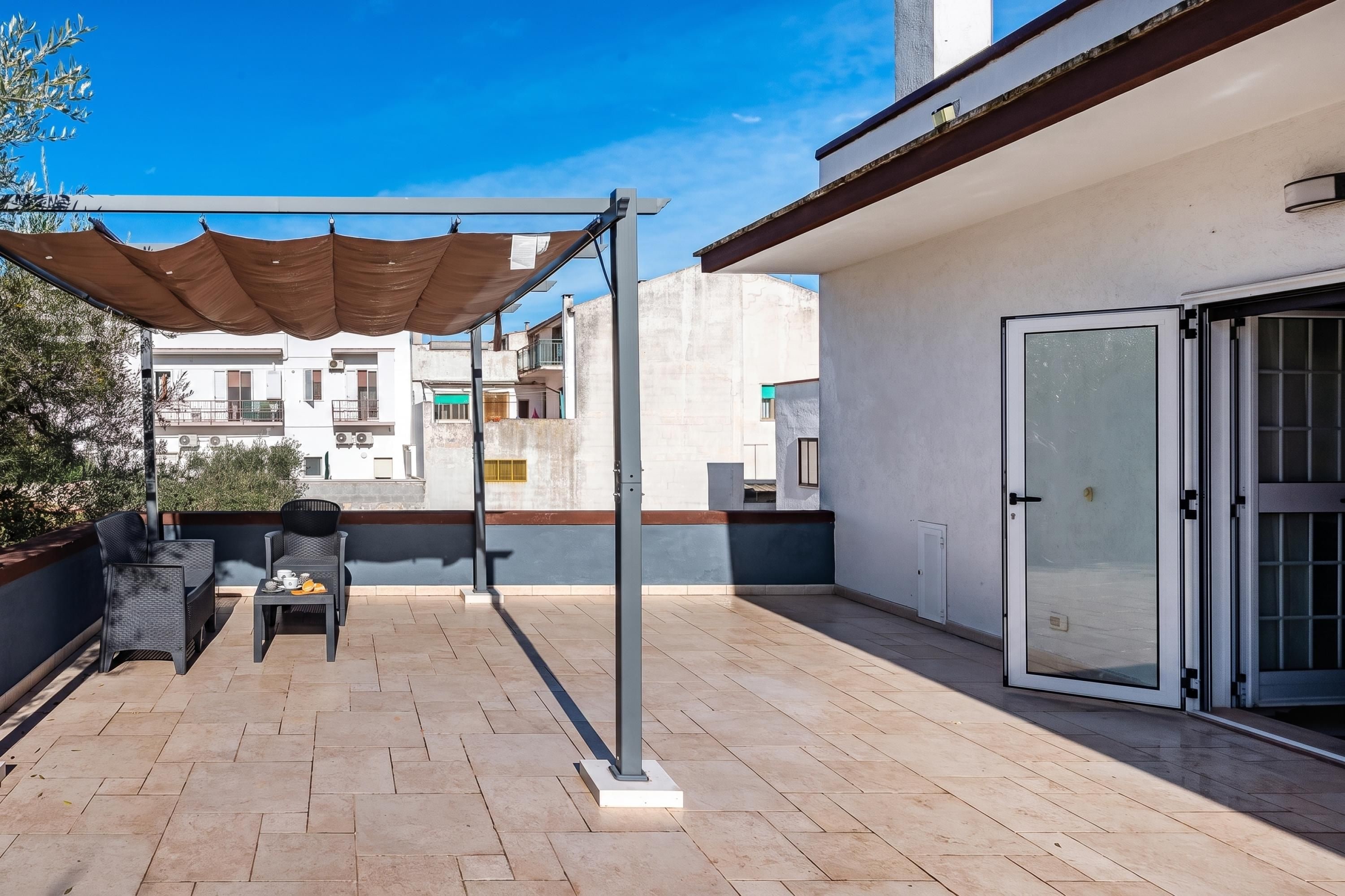 An outdoor patio area with a table and chairs, a pergola providing shade, and a view of neighboring buildings.