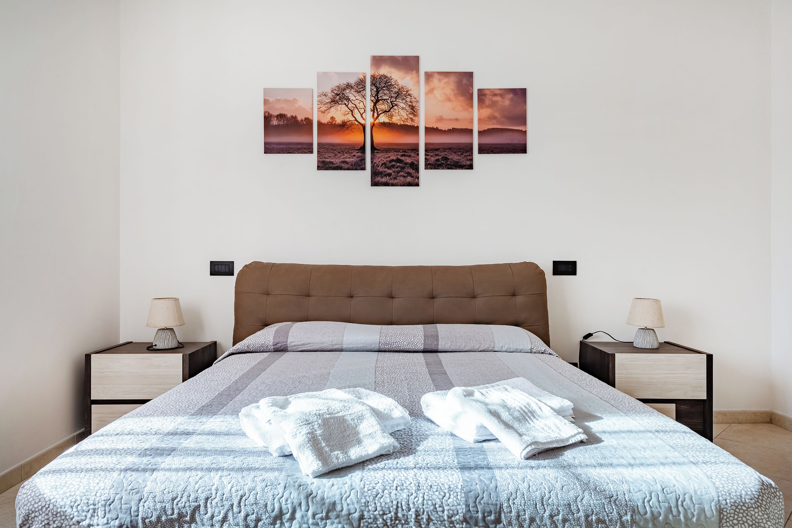 Une chambre à coucher avec un lit, deux tables de chevet, une œuvre d’art représentant un arbre accrochée au mur et une couverture sur le lit.