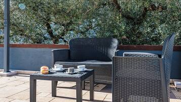 Un área de estar al aire libre con mobiliario de mimbre, una mesa con tazas y un pastel, y vistas a una vegetación densa al fondo.