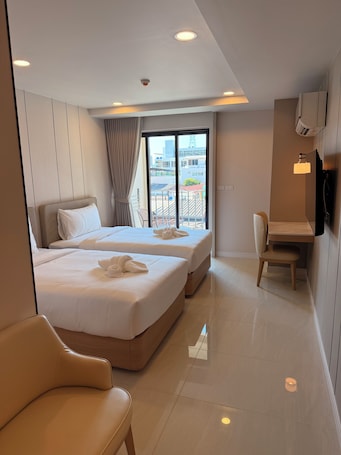 A hotel room with two beds, a desk, a chair, and a large window offering a city view.. Aston Hotel Pattaya