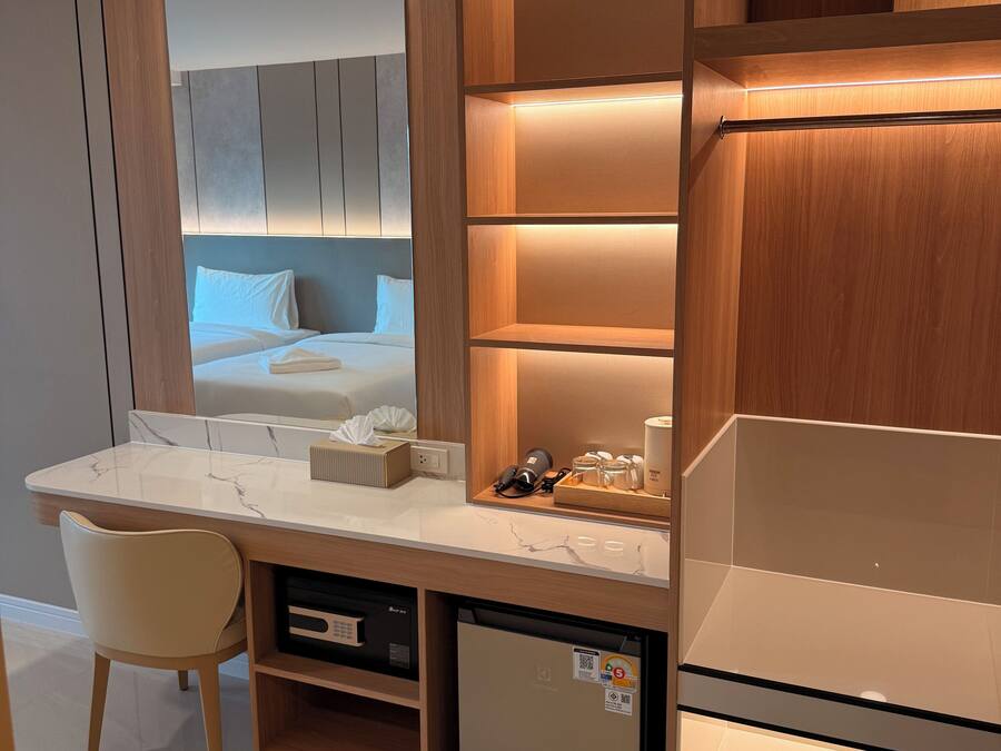 Una habitación de hotel moderna con un escritorio con mosaicos de mármol, un refrigerador compacto, un espejo integrado y una estantería de madera con iluminación integrada.