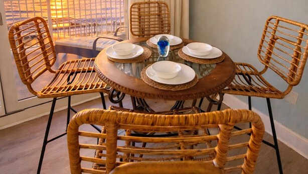 Un coin repas avec des chaises en osier et une table ronde prévue pour quatre personnes, offrant une vue sur le coucher du soleil depuis un balcon.