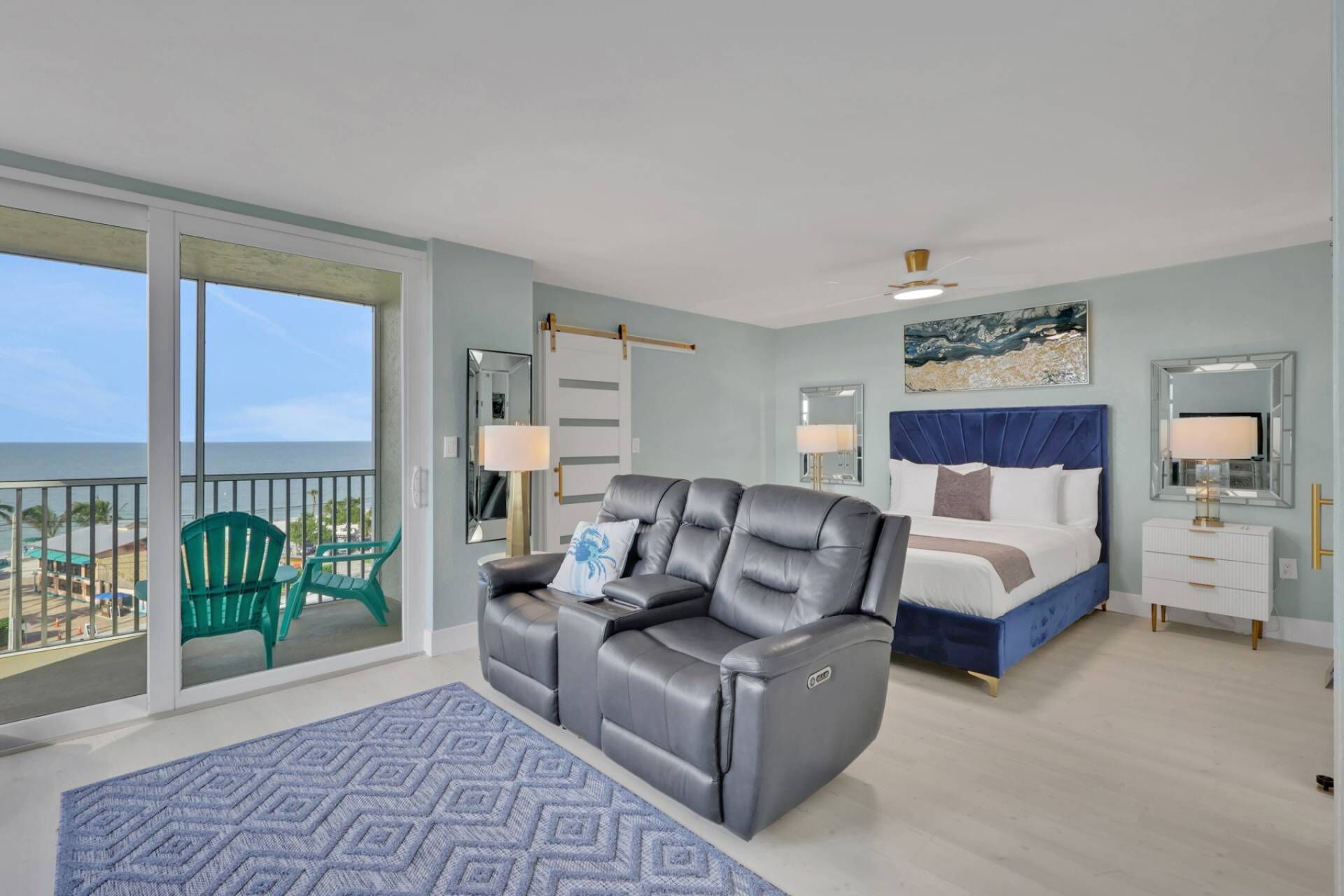 Een slaapkamer met een bed, een relaxfauteuil, een kaptafel en uitzicht op de oceaan vanaf een balkon.