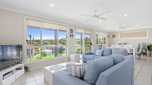 A spacious living room with a large window, a flat-screen TV, a grey sofa, and a ceiling fan. - Sea-Renity is a spacious home (Manyana)