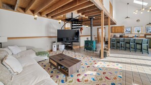 Ein geräumiger Wohnbereich mit hohen Decken, Holzbalken, einem großen Fernseher, einer Couch, einem Couchtisch und einer Küchenzeile mit Barhockern.