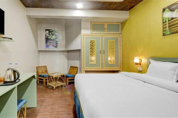 A hotel room with a bed, a desk with a kettle, two chairs, a wardrobe, and a painting on the wall. - Hi Hotel By Madras (Rishikesh)