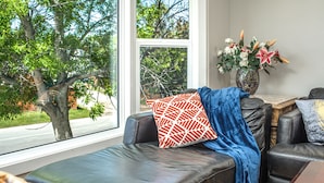 Ein Zimmer mit einer Ledersofa, einem gemusterten Kissen, einer Vase mit Blumen und Blick auf einen Baum durch ein Fenster.